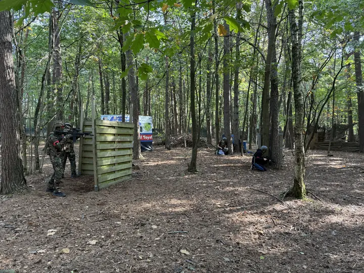 Twee spelers in camouflage leggen aan vanachter een houten schot, verderop zitten spelers in dekking
